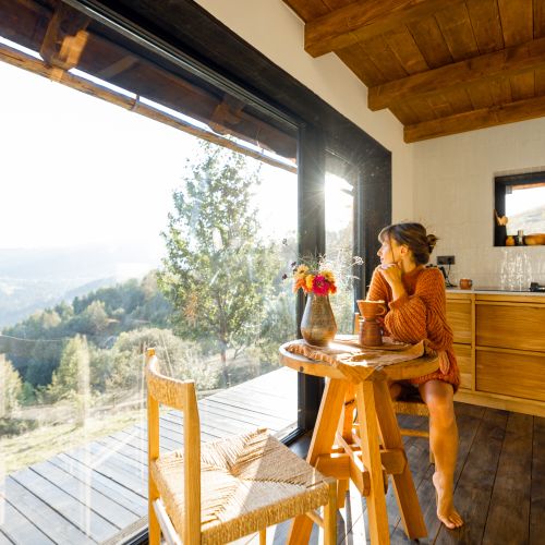 A woman in a cozy sweater enjoys her morning coffee by a large window, gazing out at the mountains. Sunlight pours into the rustic kitchen, highlighting the serene moment